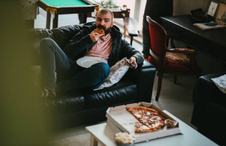 Uomo con uno stile di vita sedentario mangia una pizza sul divano